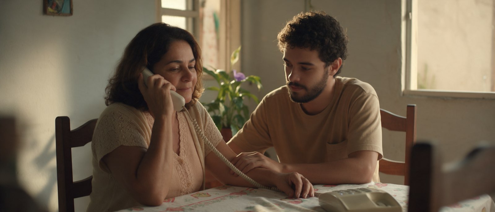 Uma mulher ao telefone à mesa da sala, ouvindo com calma, enquanto o filho adulto sentado ao lado mantém a mão sobre a dela.